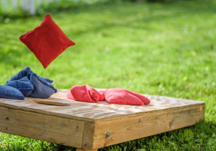 Cornhole Board on grass with four blue bags stacked on left side of hole, three red bags stacked on 